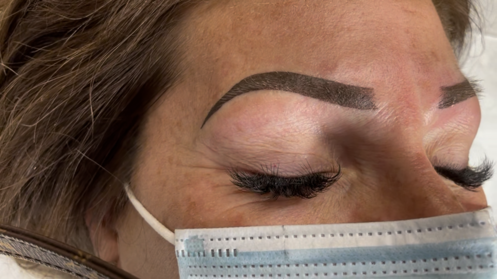 Close-up of a client's face at Just Browzin' studio in Toledo, Ohio, featuring striking, freshly defined ombre brows and luxurious volume eyelash extensions. The client is wearing a medical face mask.